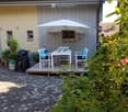 Outdoor patio area with a table, chairs, umbrella, and grill, surrounded by greenery and a paved stone pathway.