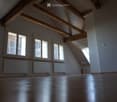 Empty attic space with wooden beams, exposed wooden ceiling, large windows, and radiators along the wall.