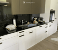Modern kitchen with white cabinets, black countertops, stainless steel sink, and various appliances including a coffee machine and oven. Wooden flooring and a white rug are visible.