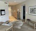 Bright, open-plan living and dining area with light wood flooring, white cabinetry, a wooden dining table with chairs, a bench, a small cabinet with a lamp, and a framed picture on the wall. A cowhide rug is placed on the floor.
