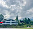 A modern two-story house with white walls, large windows, and balconies, surrounded by greenery and trees with mountains in the background.