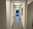 Modern hallway with white walls, grey carpeted floor, and recessed lighting. Features a bench on one side and a glass door leading to an outdoor area with a parked car visible.