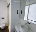 Modern bathroom with white walls, a white sink, a toilet, and a large mirror above the sink. The floor is dark wood, and there is a glass shower enclosure visible in the background.