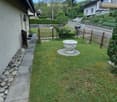 A small, well-maintained garden area with a stone pathway, a wooden fence, a decorative stone planter, and surrounding greenery.