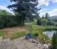Garden with a pond, small statues, potted plants, and a grassy area surrounded by trees and hedges.