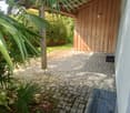 A paved outdoor area with a wooden shed, surrounded by lush greenery and palm leaves. The ground is covered with interlocking stone tiles, and there is a metal grate near the wall.
