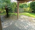 A covered outdoor area with a stone-paved floor, wooden beams, and a lush garden with palm trees and greenery.
