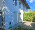 Two-story house with white exterior, multiple windows, and a small balcony on the upper level. The house has a paved pathway leading to the entrance and a garden area with greenery.