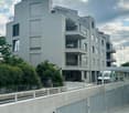 Modern multi-story residential building with grey facade, multiple balconies, and large windows. Features a raised ground floor with a concrete barrier and parking area.