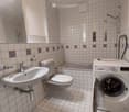 Bathroom with white tiles, a sink, toilet, bathtub, and a washing machine. Features a shower area with a glass enclosure and a mirror above the sink.