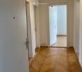 Empty hallway with wooden herringbone flooring, white walls, and open doorway leading to another room.