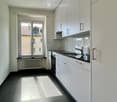 Modern, unfurnished kitchen with white cabinets, stainless steel sink, and a large window offering a view of a neighboring building. The space features a dark tiled floor and a sleek, minimalist design.