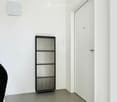 Empty room with white walls, grey carpeted floor, and a closed door with a glass panel. There is a small shelf with a decorative item on the left wall.