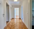 Clean, modern hallway with herringbone wooden flooring, white walls, and recessed lighting. Doors lead to adjacent rooms.