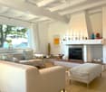 Bright living room with white walls, wooden floor, large window with view, fireplace, sofa, ottoman, coffee table, decorative items on mantel.