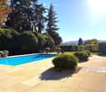 A well-maintained garden area with a swimming pool, surrounded by lush greenery, trees, and neatly arranged potted plants. The pool is surrounded by a tiled patio area, and the background features a scenic view of hills and trees.