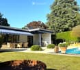 Well-maintained garden with a swimming pool, patio furniture, and a modern house with a striped awning.