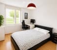 Modern bedroom with a double bed, nightstand, desk with a computer, and a large window with blinds. The room features a wooden floor and white walls.