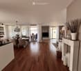 Open-plan living and dining area with wooden flooring, large windows, dining table with chairs, TV, cabinet, decorative vases, and a chandelier.