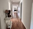 A long, well-lit hallway with wooden flooring, white walls, and several doors on the right side. A small table with a lamp and a basket is placed on the left side.