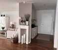 A well-lit living room with a white fireplace, a sofa, coffee table, table lamps, and decorative vases. The room features wooden flooring and a hallway leading to another area.