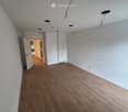 Empty room with light-colored walls, wooden flooring, and several ceiling spotlights. An open doorway leads to another room with similar flooring.