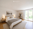Modern bedroom with a large bed, nightstands, lamps, artwork, clothing rack, and plants.