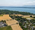 Aerial view of a suburban area with houses, fields, and a large body of water in the background. The area includes residential buildings, agricultural fields, and a waterfront with boats.
