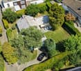 Aerial view of a residential house with a well-maintained garden, driveway, and surrounding greenery. The house features a tiled roof and is surrounded by trees and shrubs.
