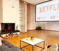 Modern living room with wooden flooring, a large screen displaying streaming services, a fireplace with stacked firewood, a sofa, and a coffee table with a decorative bowl and remote controls.