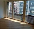 Empty room with wooden flooring and large glass doors leading to a balcony, showing a cityscape view.