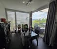 Modern dining area with a glass dining table, chairs, a cabinet with books and decorative items, and a view of a balcony with outdoor furniture.