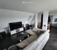 Modern living room with a white sectional sofa, coffee table, large flat-screen TV, speakers, and a fish tank. The room features dark flooring and white walls with a skylight window.