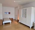 A well-lit bedroom with a white bed, a large wardrobe with glass doors, a nightstand, and a wicker basket. The room features light-colored walls and a wooden floor.