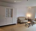 A well-lit living room with a patterned sofa, matching armchair, glass coffee table, and a large white wardrobe with glass-paneled doors.