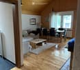 Bright, open living and dining area with wooden floors, a slanted wooden ceiling, and large windows. Features a sofa, coffee table, dining table with chairs, and a lamp.