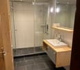 Modern bathroom with dark tiled floor, white sink, wooden cabinet, large mirror, and glass-enclosed shower area.