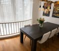 Dining room with a dark wooden dining table, four light-colored chairs, a vase with flowers, and large windows with white blinds.