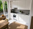 Bright, modern kitchen with white cabinets, a central island, and a dining area. Features include a wooden dining table, a white oven, a sink with a modern faucet, and decorative items like vases and bottles.