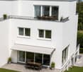 Modern two-story white house with large windows, a covered patio, and a rooftop terrace with outdoor furniture.