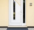Front entrance door with a modern white frame and glass panels, featuring a decorative orange diagonal design on the glass. A black doormat is placed on the gray tiled ground in front of the door.