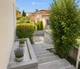 A modern garden area with a paved pathway, potted plants, and surrounding greenery, leading to a grassy lawn and neighboring houses.