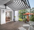 Wooden deck with white wicker chairs, small table, outdoor umbrella, and a large ceramic vase. The deck is covered by a black and white striped awning, with a view of a garden and trees.