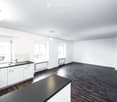 Empty room with white walls, dark wooden flooring, and a small kitchen area featuring white cabinets and a sink.