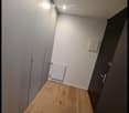 A narrow hallway with wooden flooring, white walls, and a radiator on one side. The hallway has a closed door at the end and a light fixture on the ceiling.