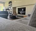 Modern living room with a grey sectional sofa, ottoman, armchair, coffee table, flat-screen TV, bookshelf, laptop, fireplace, and wood stack.