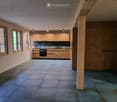 Modern kitchen with light wooden cabinets, stainless steel oven, and tiled floor. Large windows provide natural light.