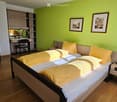 Bright bedroom with green walls, double bed with yellow bedding, nightstand, desk with chair, and framed pictures on the wall.