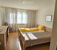 Cozy bedroom with two single beds, a desk with a laptop, a chair, and a television mounted on the wall. The room features wooden flooring and large windows with curtains.
