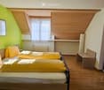 Bright bedroom with two single beds, wooden floor, slanted wooden ceiling, and a TV area with a chair and table.